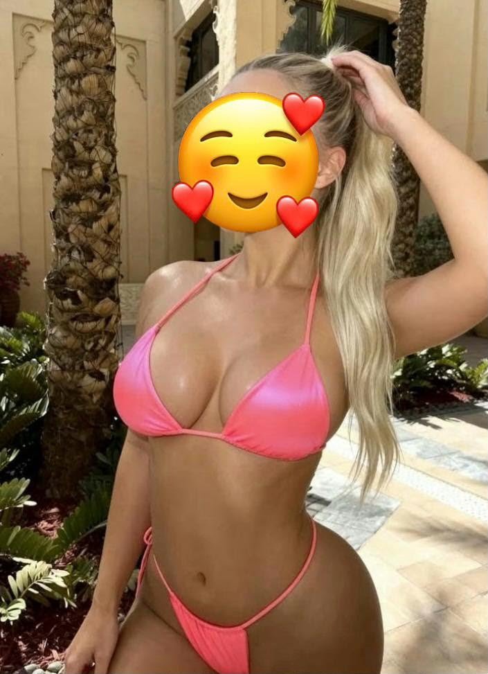 Woman in a pink bikini posing poolside among palm trees; face covered by a heart-eyed smiling emoji.