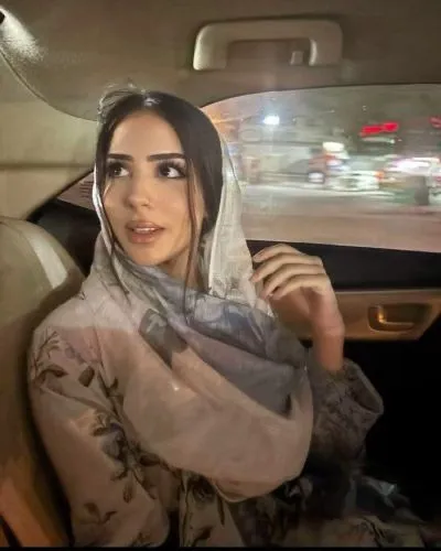 Woman in a light floral hijab sitting in the back seat of a car, looking toward the passenger side as city lights pass by outside the window.