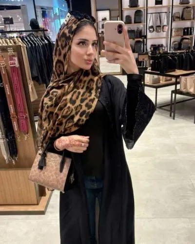 Woman in a leopard-print hijab and black outfit takes a selfie in a store with handbags in the background.